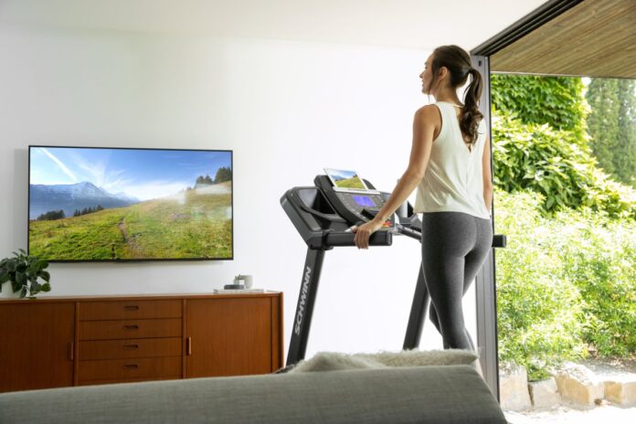 Kobieta ćwiczy w domu cardio na bieżni w trybie marszu, oglądając trasę na ekranie