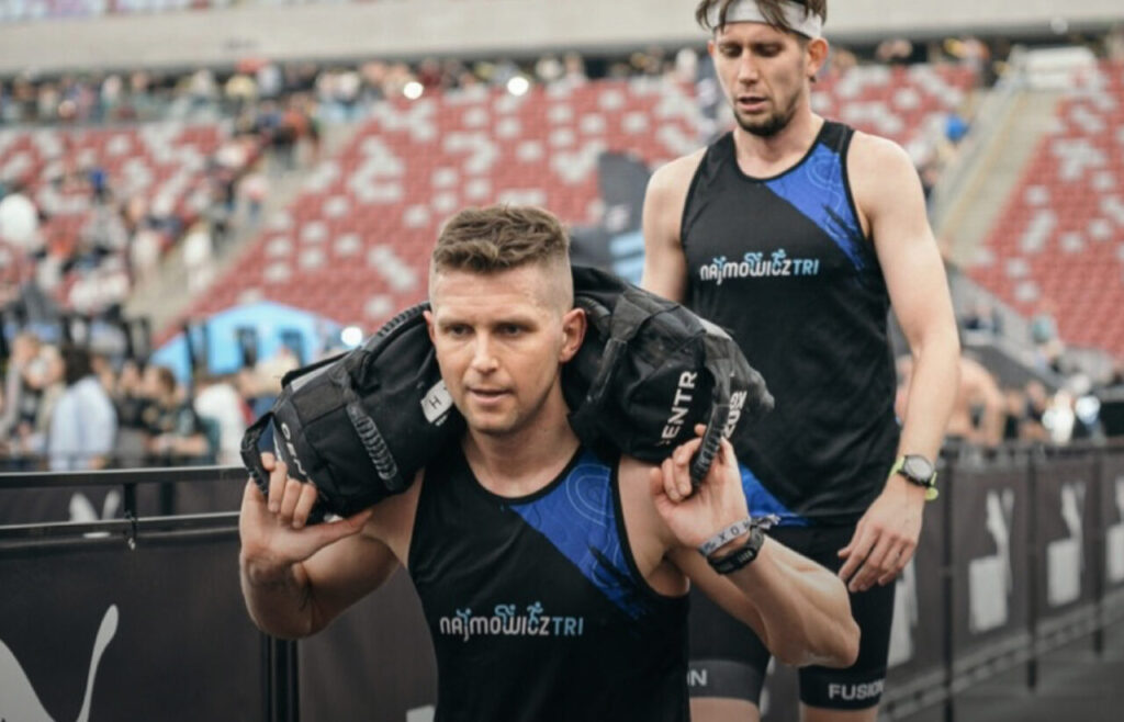 Zawodnicy podczas zawodów HYROX wykonujący wykroki z workiem z piaskiem w trakcie wyścigu łączącego bieganie i ćwiczenia siłowe