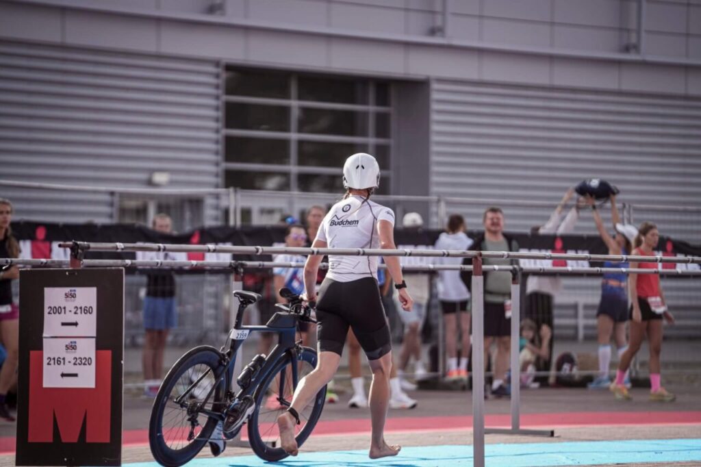Zawodniczka IRONMAN prowadzi rower w strefie zmian T1 podczas zawodów triathlonowych
